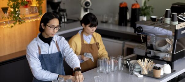 Two baristas stand behind a café counter; one arranges glassware on a tray while the other dries a glass with a towel, showcasing Office Beverage Solutions Singapore for modern workplace refreshments.